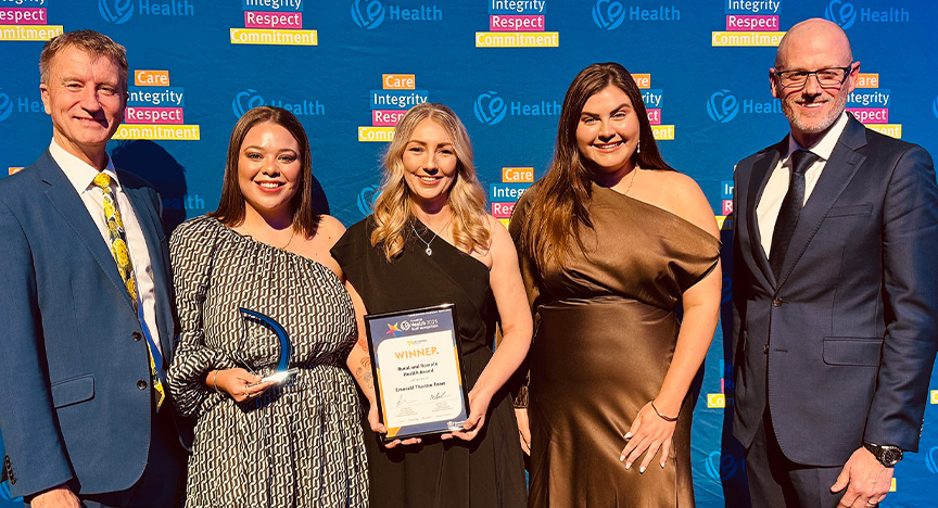 Healthcare workers wearing cocktail attire stand in front of a royal blue media wall printed with Central Queensland Health values at an awards presentation evening at a conference centre.