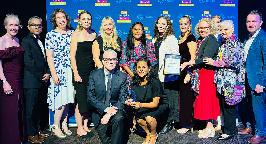 Healthcare workers wearing cocktail attire stand in front of a royal blue media wall printed with Central Queensland Health values at an awards presentation evening at a conference centre.