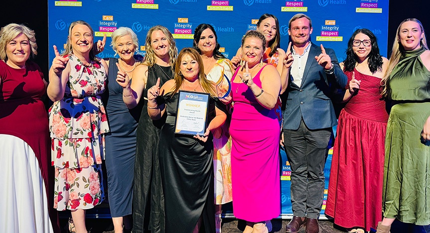 Healthcare workers wearing cocktail attire stand in front of a royal blue media wall printed with Central Queensland Health values at an awards presentation evening at a conference centre.