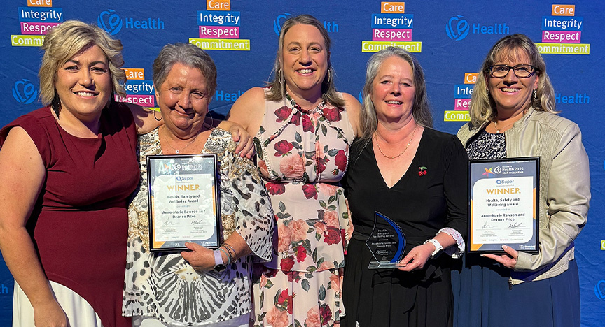 Healthcare workers wearing cocktail attire stand in front of a royal blue media wall printed with Central Queensland Health values at an awards presentation evening at a conference centre.